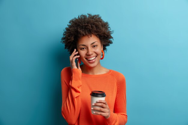 Alegre y despreocupada chica milenaria étnica tiene una conversación telefónica agradable, mantiene el teléfono inteligente cerca del oído, tiene una linda sonrisa y un estado de ánimo feliz, bebe bebidas aromáticas, usa un jersey naranja, aislado en azul