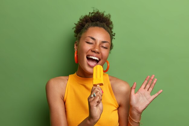 Alegre y despreocupada mujer afroamericana canta en un helado amarillo como en un micrófono, disfruta de un delicioso producto de verano, está muy feliz, aislada sobre una pared verde vívida, levanta la mano, muestra dientes blancos