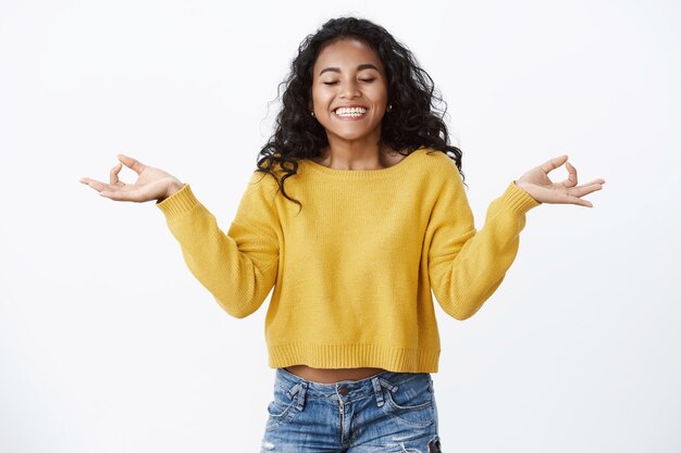 Alegre despreocupada optimista, chica con cabello rizado, ojos cerrados, inhala aire fresco, sonriendo aliviada y libre, meditando con las manos extendidas hacia los lados, signos de mudra zen, pared blanca