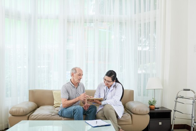 Alegre doctora asiática visitando pacientes varones caucásicos senior en casa