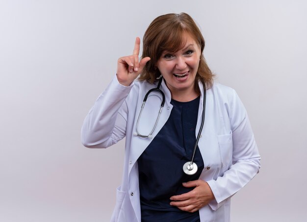 Alegre doctora de mediana edad con bata médica y un estetoscopio apuntando con el dedo hacia arriba y poniendo la mano en el vientre en la pared blanca aislada con espacio de copia