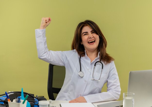 Alegre doctora de mediana edad con bata médica con estetoscopio sentado en el escritorio, trabajo en una computadora portátil con herramientas médicas, haciendo un gesto fuerte en una pared verde aislada con espacio de copia