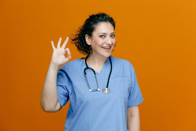 Alegre doctora de mediana edad con uniforme y estetoscopio alrededor del cuello mirando a la cámara mostrando el signo de ok aislado en el fondo naranja