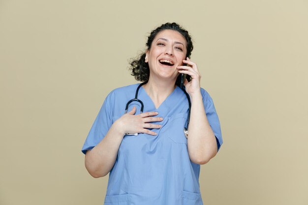 Alegre doctora de mediana edad con uniforme y estetoscopio alrededor del cuello mirando a un lado hablando por teléfono riéndose mientras mantiene la mano en el pecho aislada en un fondo verde oliva