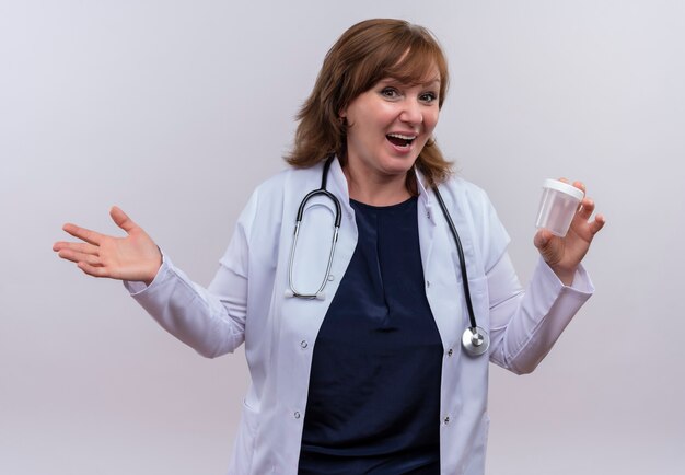 Alegre doctora de mediana edad vistiendo bata médica y estetoscopio sosteniendo vaso médico en pared blanca aislada
