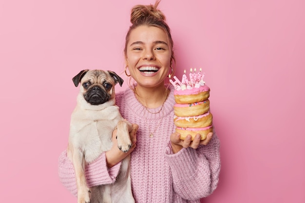 La alegre dueña de una mascota posa con un perro pug de pedigrí y donas contentas de celebrar el cumpleaños de un animal doméstico viste un suéter de punto aislado sobre fondo rosa Concepto de celebraciones felices