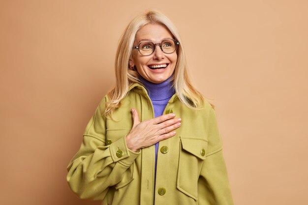 Alegre y elegante mujer rubia de cuarenta años vestida con ropa fashioable mantiene la mano en el pecho y sonríe positivamente al recordar que algo gracioso sucedió con ella.