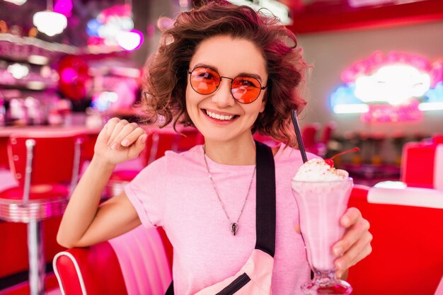 Alegre y elegante mujer sonriente en un colorido café retro vintage americano de los años 50 sentado en la mesa bebiendo un cóctel de batido de leche con gafas de sol rosas divirtiéndose