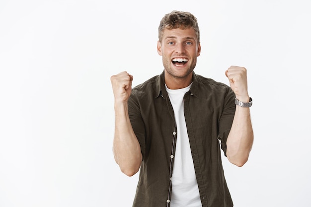 Alegre emocionado feliz atractivo rubio con dientes blancos y cerdas levantando los puños con alegría, sonriendo satisfecho triunfando, celebrando la victoria o el éxito, reaccionando a noticias impresionantes sobre la pared gris.