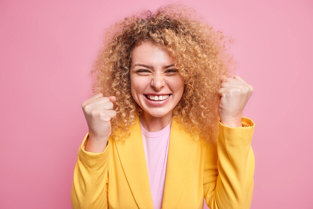 Alegre empresaria de pelo rizado llena de alegría aprieta los puños como el campeón celebra la victoria oye una excelente noticia vestida con chaqueta amarilla aislada sobre una pared rosa. Mujer corporativa triunfante