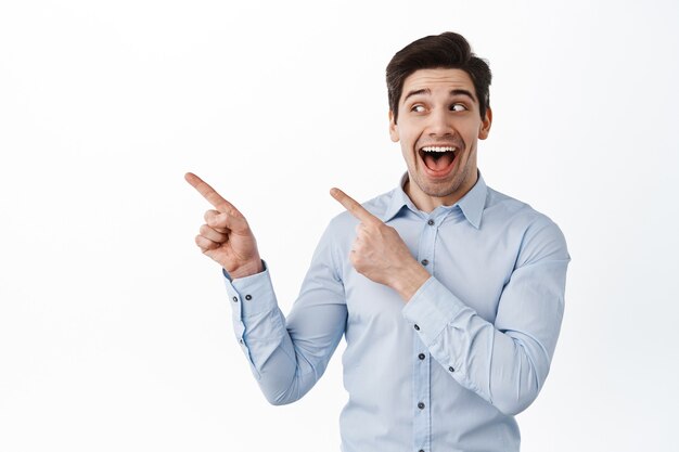 Alegre empresario jadeando fascinado, mirando y apuntando a un lado a la izquierda, mostrando publicidad, de pie sobre una pared blanca con camisa de cuello azul