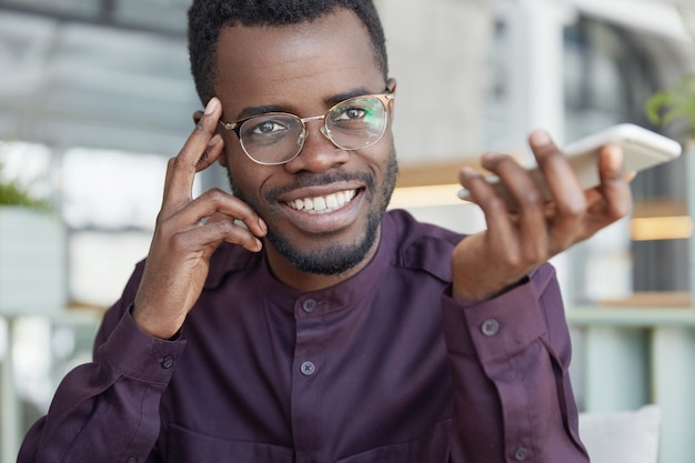 Alegre empresario próspero de piel oscura con gafas, tiene una amplia sonrisa brillante, sostiene un teléfono inteligente y confirma la banca en la aplicación