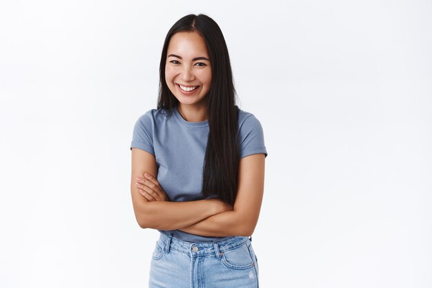 Alegre y encantadora mujer asiática en camiseta, jeans riendo y de pie con entusiasmo sobre la pared blanca, cruzando las manos sobre el pecho sonriendo modestamente, pasando el rato con nuevos compañeros de trabajo durante la fiesta