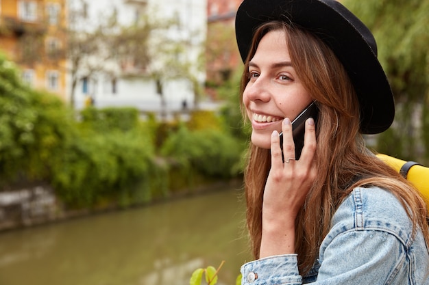 Alegre y encantadora turista se encuentra contra el fondo de un río pequeño en la ciudad, tiene conversación por teléfono móvil, utiliza conexión de roaming