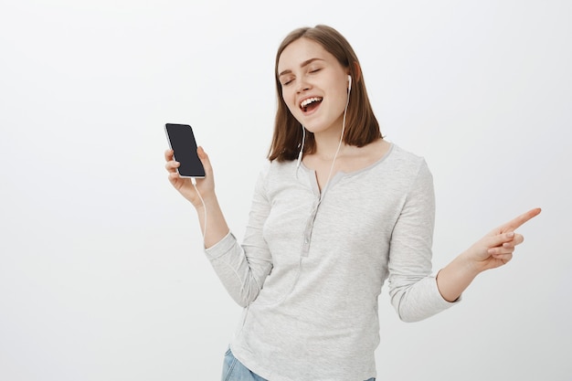 Alegre y entusiasta mujer europea guapa con corte de pelo marrón cerrando los ojos alejándose de la diversión y la alegría mientras escucha música en auriculares sosteniendo un teléfono inteligente sobre una pared gris