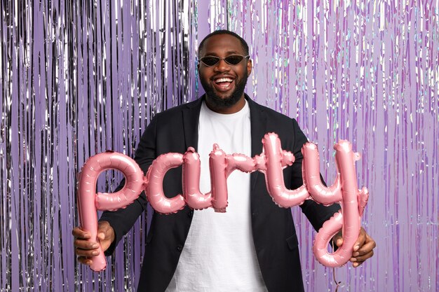 Alegre estudiante afroamericano graduado tiene una fiesta con compañeros de grupo después de salir de la universidad, usa elegantes lentes de sol, traje formal, sostiene un globo en forma de letras, se para sobre una cortina de oropel