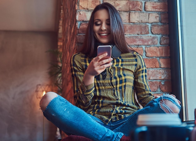 Foto gratuita alegre estudiante morena con ropa informal escuchando música mientras usa un smartphone mientras se sienta en el alféizar de una ventana en una habitación con un interior de loft.