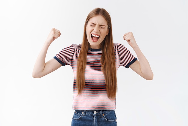 Alegre, exitosa y atractiva chica con camiseta a rayas hizo que los sueños se hicieran realidad finalmente logrando el objetivo, levantando las manos en señal de triunfo, gritando encantada de lograr la victoria, sintiéndose como un campeón