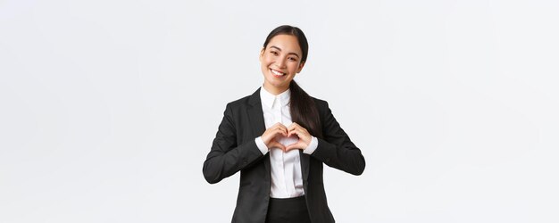 Una alegre y exitosa mujer de negocios asiática con traje negro cuida a sus clientes mostrando un gesto de corazón y sonriendo feliz brindando un servicio cariñoso a los clientes de fondo blanco