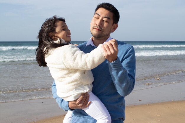 Alegre familia asiática caminando juntos en la playa. Padre e hija con ropa informal bailando cerca del agua y riendo. Unión, amor, concepto de crianza