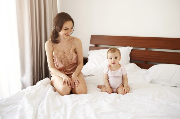 Alegre feliz mamá joven en ropa de dormir y su pequeña hija sonriente jugando sentado en la cama por la mañana.