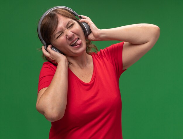 Alegre feliz mujer de mediana edad en camiseta roja con auriculares disfrutando de su música favorita sacando la lengua de pie sobre la pared verde