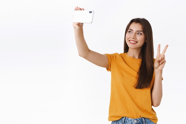 Alegre, feliz y saliente linda bloguera graba un video durante un viaje por Europa levantando un teléfono inteligente posando para una selfie con un signo de paz o victoria de fondo blanco Concepto de tecnología