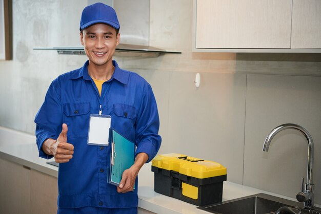 Alegre fontanero asiático de pie cerca del fregadero de la cocina y mostrando el pulgar hacia arriba