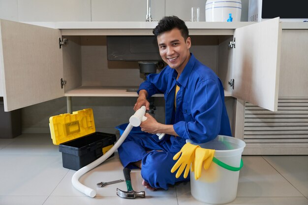 Alegre fontanero asiático sentado en el piso y reparando el fregadero de la cocina