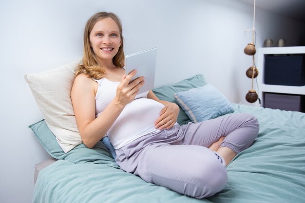 Alegre futura mamá con tableta en el dormitorio