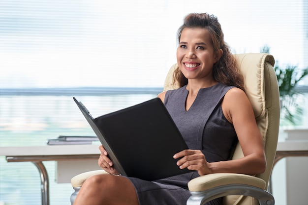 Alegre gerente de recursos humanos con carpeta de falta sentado en la entrevista de trabajo y mirando hacia los lados