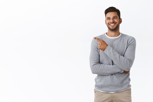 Alegre, guapo, amable, barbudo, con un suéter gris, riendo de una broma divertida o un anuncio, sonriendo despreocupado y encantado, charlando con un amigo y apuntando a la izquierda, pared blanca