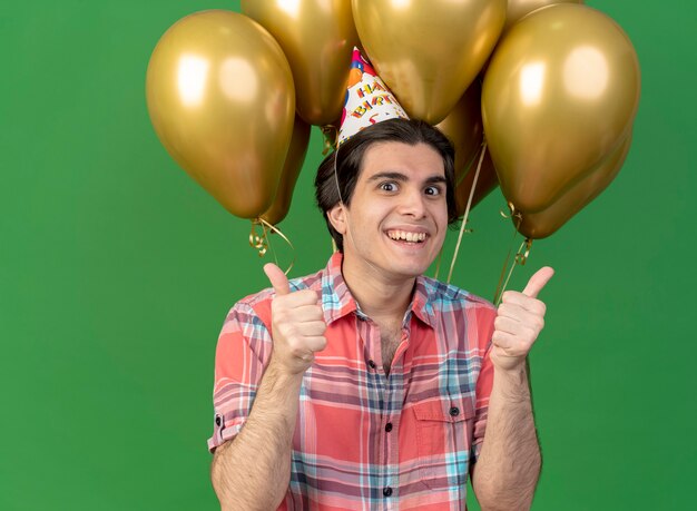 Alegre guapo caucásico hombre vestido con gorro de cumpleaños se encuentra delante de globos de helio Thumbs up con dos manos