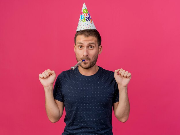 Foto gratuita alegre guapo caucásico hombre vestido con gorro de cumpleaños mantiene el puño soplando el silbato del partido aislado sobre fondo rosa con espacio de copia