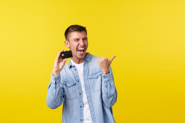 Alegre guapo rubio apuntando con el dedo a la derecha y mostrando la tarjeta de crédito, riendo de alegría, anunciando servicios bancarios, excelentes precios en la tienda, yendo de compras, de pie con fondo amarillo.