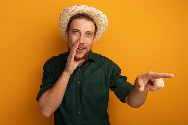 Alegre guapo rubio con sombrero de playa sostiene la mano cerca de la boca y apunta al lado aislado en la pared naranja