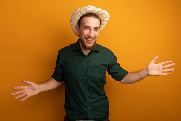 Alegre guapo rubio con sombrero de playa tiene las manos abiertas aisladas en la pared naranja