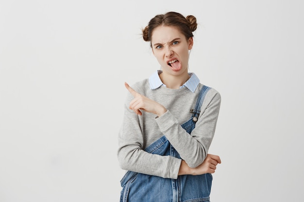 Alegre hembra adulta que muestra el dedo sobre la pared blanca. Mujer segura con doble bollos sacando la lengua y expresando que no ama su elección. Copia espacio