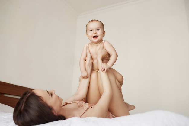 Alegre hermosa mamá y su bebé jugando sonriendo regocijo acostado en la cama en su casa