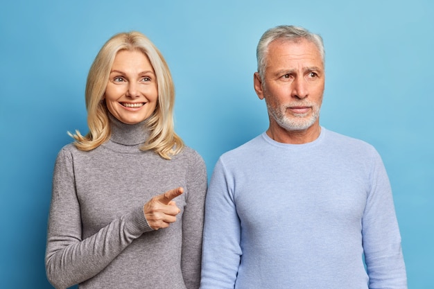 Alegre hermosa mujer de mediana edad indica el dedo índice en la distancia y muestra algo al esposo que está concentrado en algún lugar con expresión seria