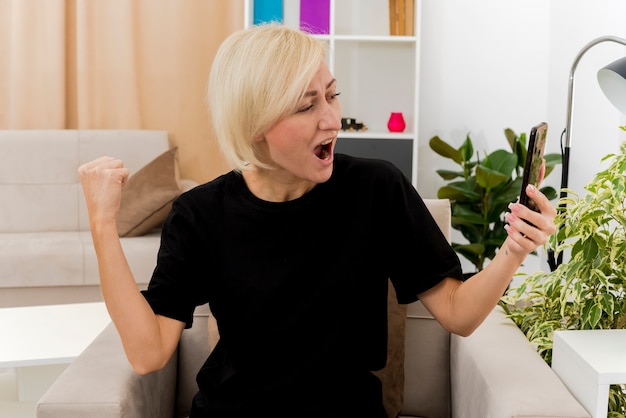 Alegre hermosa mujer rusa rubia sentada en un sillón manteniendo el puño hacia arriba y mirando el teléfono dentro de la sala de estar