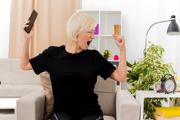 Alegre hermosa mujer rusa rubia sentada en un sillón manteniendo el puño y sosteniendo el teléfono mirando al costado dentro de la sala de estar