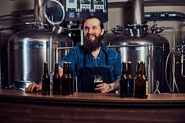 Un alegre hipster barbudo tatuado con una camisa de jeans y un delantal que trabaja en una fábrica de cerveza, parado detrás de un mostrador, sostiene un vaso de cerveza para el control de calidad.