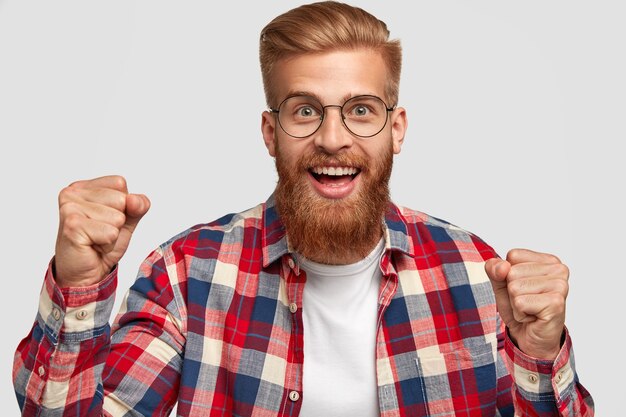 Alegre hipster con expresión divertida, aprieta los puños, celebra un día exitoso, tiene un peinado moderno y una barba pelirroja, viste una camisa a cuadros brillante, aislada sobre una pared blanca. Concepto de triunfo