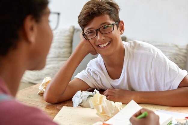 Alegre hipster masculino asiático usa gafas ópticas, camiseta blanca informal, responde preguntas durante la entrevista, el periodista irreconocible se sienta, escribe notas en el bloc de notas. Los adolescentes trabajan juntos