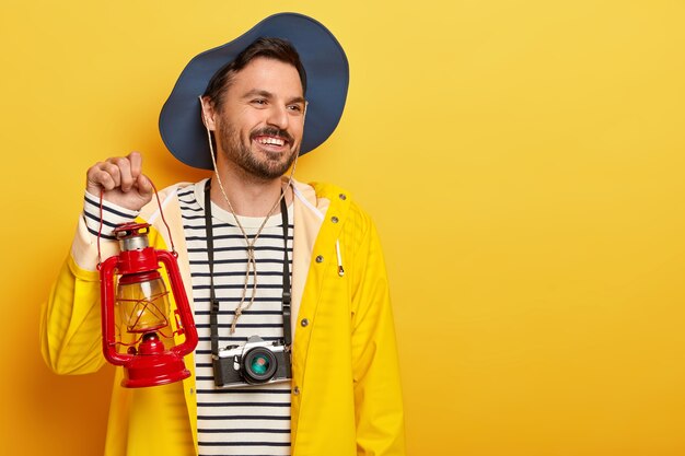 Alegre hombre activo sostiene una lámpara de gas, camina en el bosque, usa una cámara retro en el cuello, mira con una sonrisa a un lado, usa sombrero e impermeable, posa sobre una pared amarilla