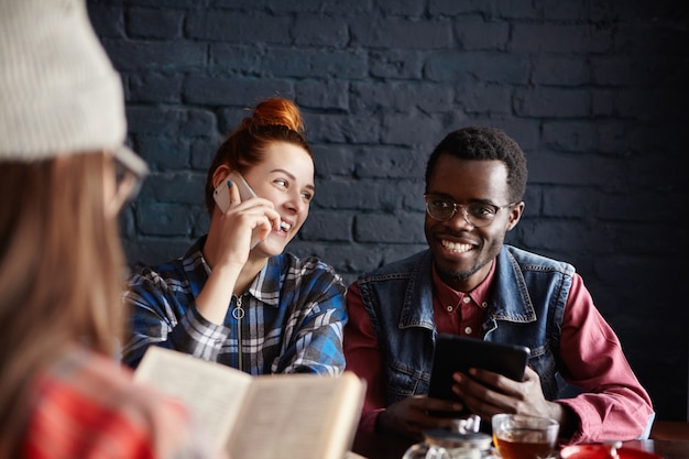 Alegre hombre africano con tableta digital cenando en el café junto con sus dos amigas: despreocupada mujer de jengibre teniendo una agradable conversación por teléfono móvil