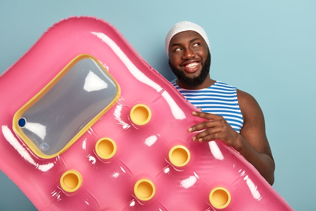 Alegre hombre afro feliz con una brillante sonrisa con dientes, sostiene un colchón inflable, listo para relajarse durante el día soleado en la playa