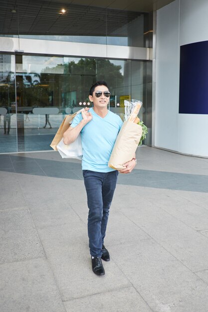 Alegre hombre asiático posando con bolsas de compras y comestibles en la entrada del edificio