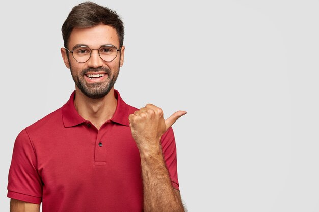 Alegre hombre caucásico barbudo con sonrisa suave, vestido con ropa informal, le muestra la dirección al lugar agradable, indica con el pulgar a un lado